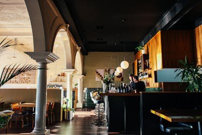 The interior of a stylish bar or restaurant featuring a dark bar counter with bottles, pendant lights, a textured ceiling, and people gathered around the bar, with architectural arches on the left.