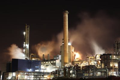 A large industrial complex with multiple towering structures, chimneys, and numerous pipes, brightly lit and emitting smoke and steam against a dark night sky.