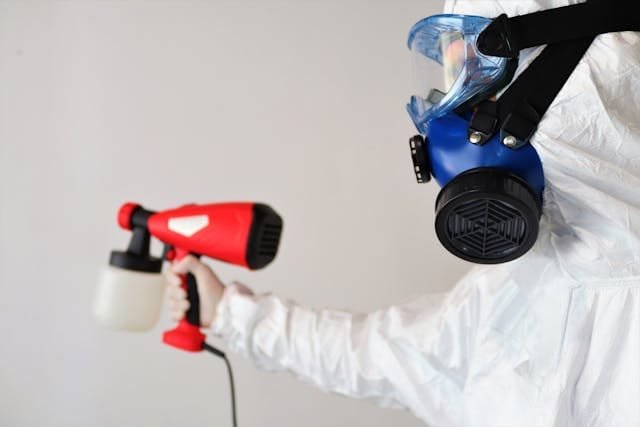 A person wearing a full white hazmat suit, blue and black respirator mask, and safety goggles, holding a red and black paint sprayer.