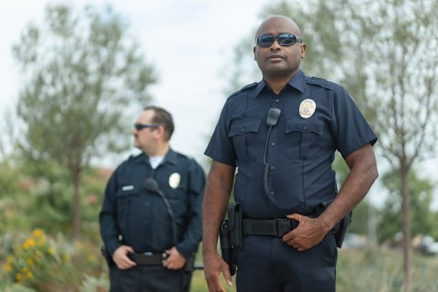 Two security guards in dark blue uniforms and sunglasses standing outdoors, with one in the foreground looking directly at the viewer and the other in the background looking to the side.