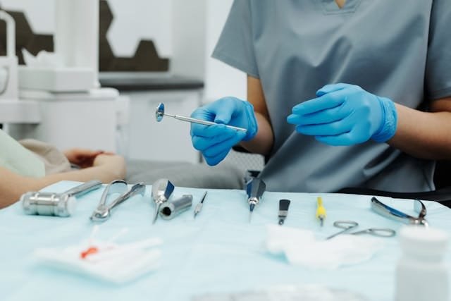 A healthcare professional wearing blue gloves and grey scrubs, holding a dental mirror above a tray of medical instruments.