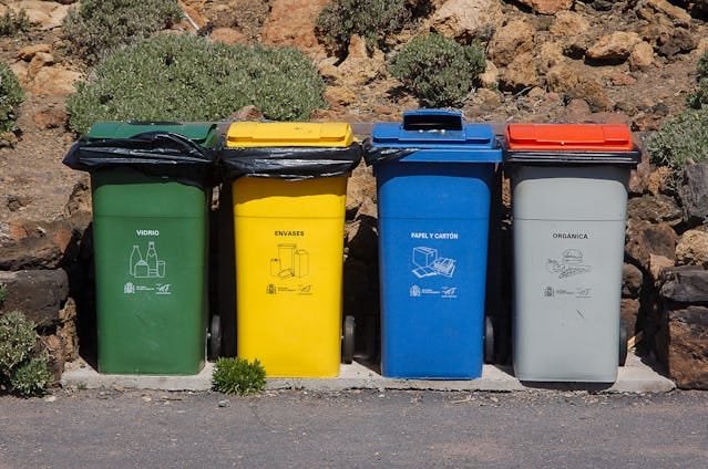 Four different colored recycling bins (green for glass, yellow for packaging, blue for paper and cardboard, and grey for organic) lined up outdoors against a rocky, vegetated slope.