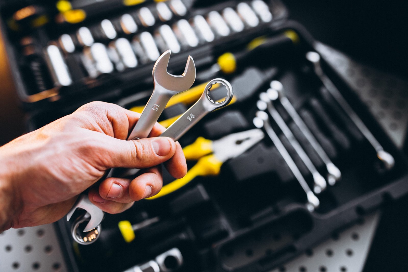A hand holding two open-end wrenches over an open toolbox filled with various mechanical tools, suggesting readiness for repair or maintenance tasks.