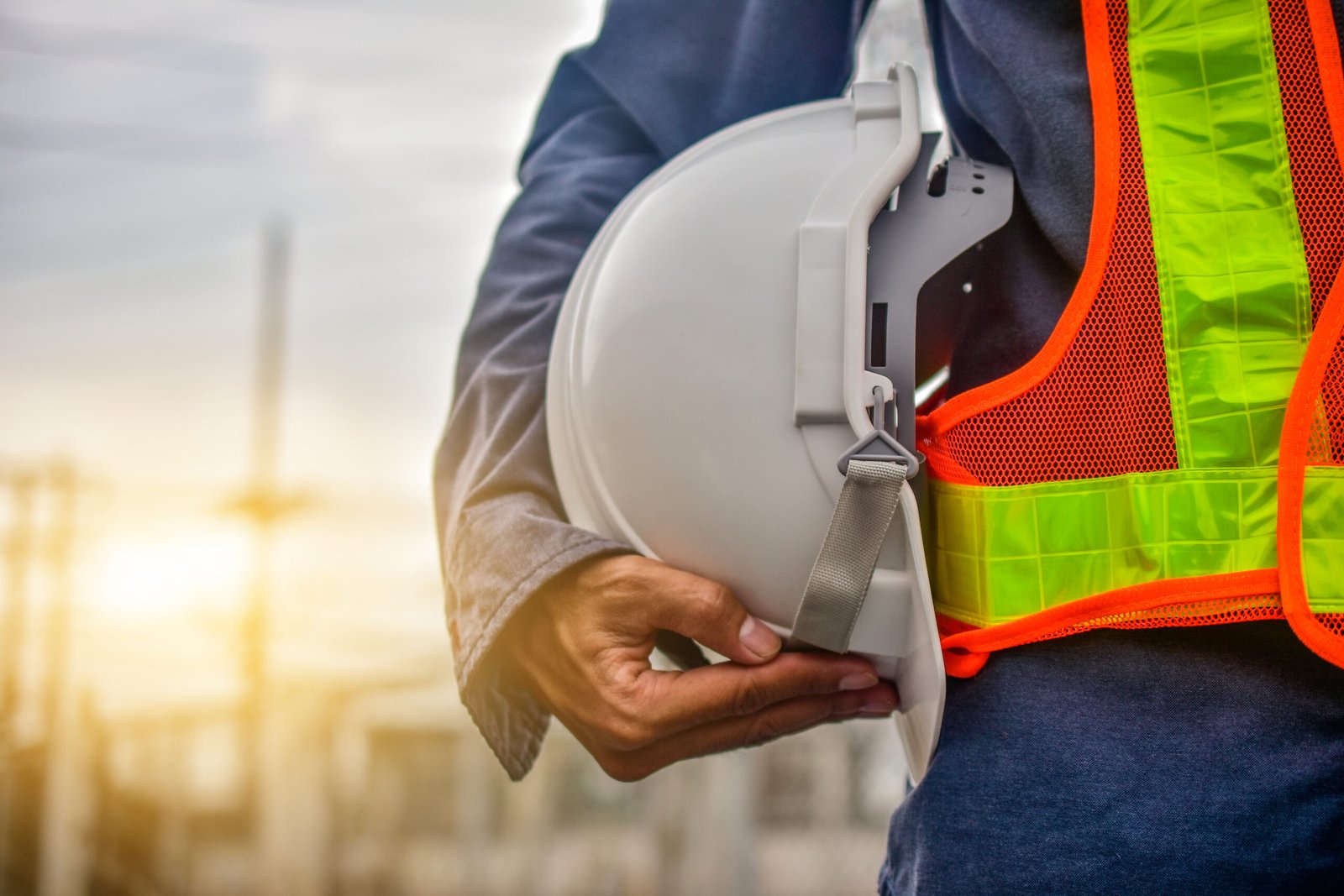Close-up of a construction worker, wearing a high-visibility vest, holding a white hard hat with their hand. Sunlight flares in the background.