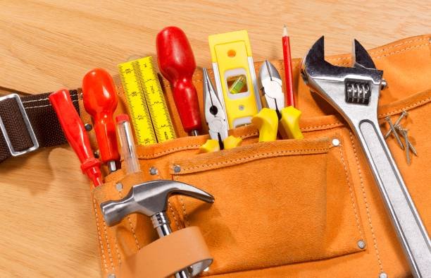A close-up of a brown leather tool belt filled with various tools including a hammer, wrench, pliers, screwdriver, level, and pencil, all resting on a wooden surface.