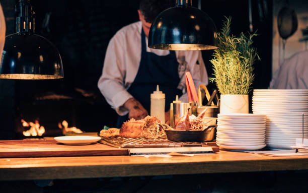 A chef in a white uniform and dark apron works at a kitchen counter with food on a grill, stacks of plates, and herbs under warm lighting.