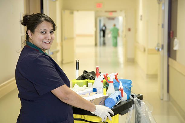 A smiling female environmental services worker, possibly of South Asian descent, wearing scrubs and white gloves, pushes a cleaning cart down a brightly lit hospital hallway.