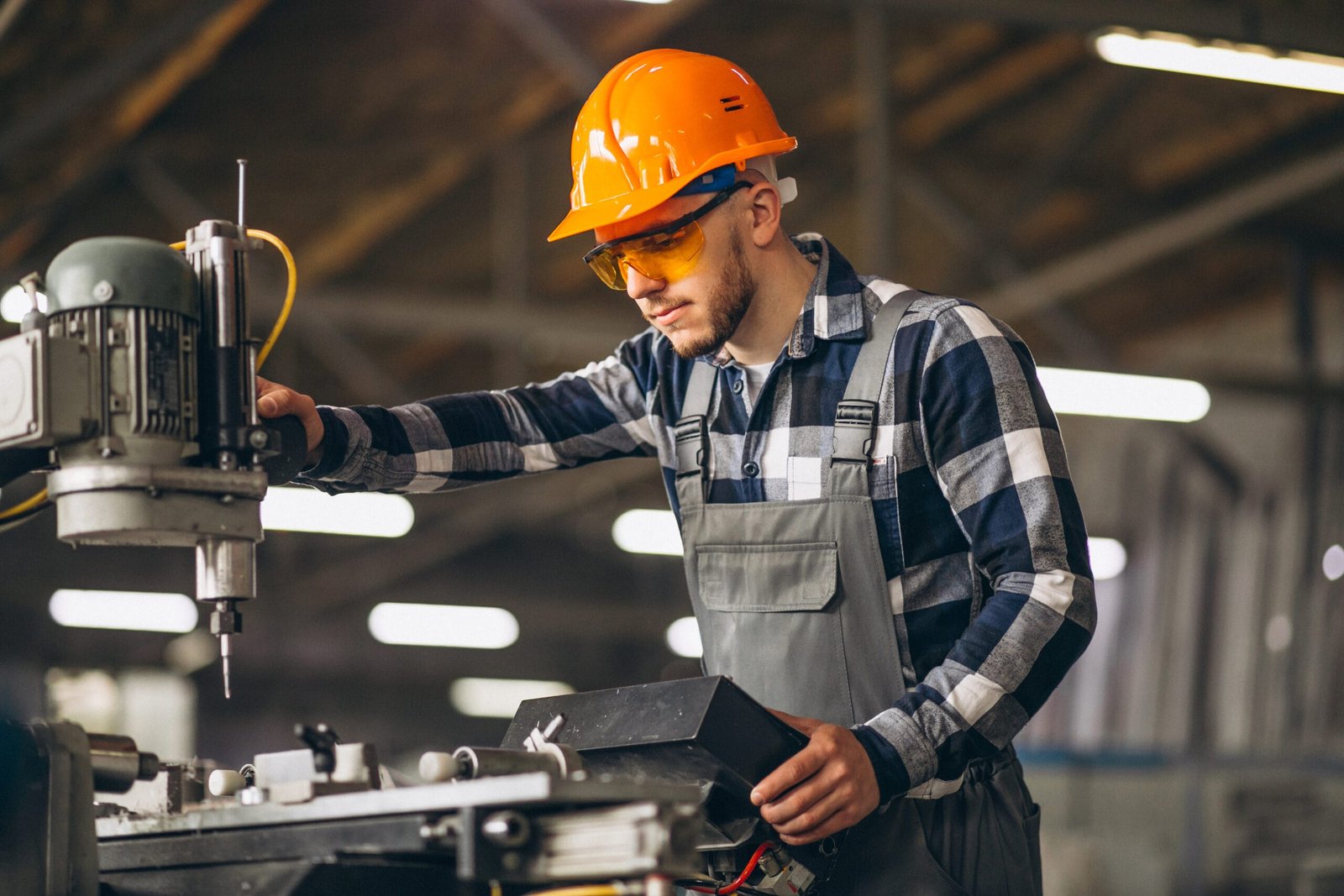 A male technician or engineer wearing an orange hard hat, safety glasses, and plaid shirt over work overalls, intently operating machinery in what appears to be a factory or workshop setting.
