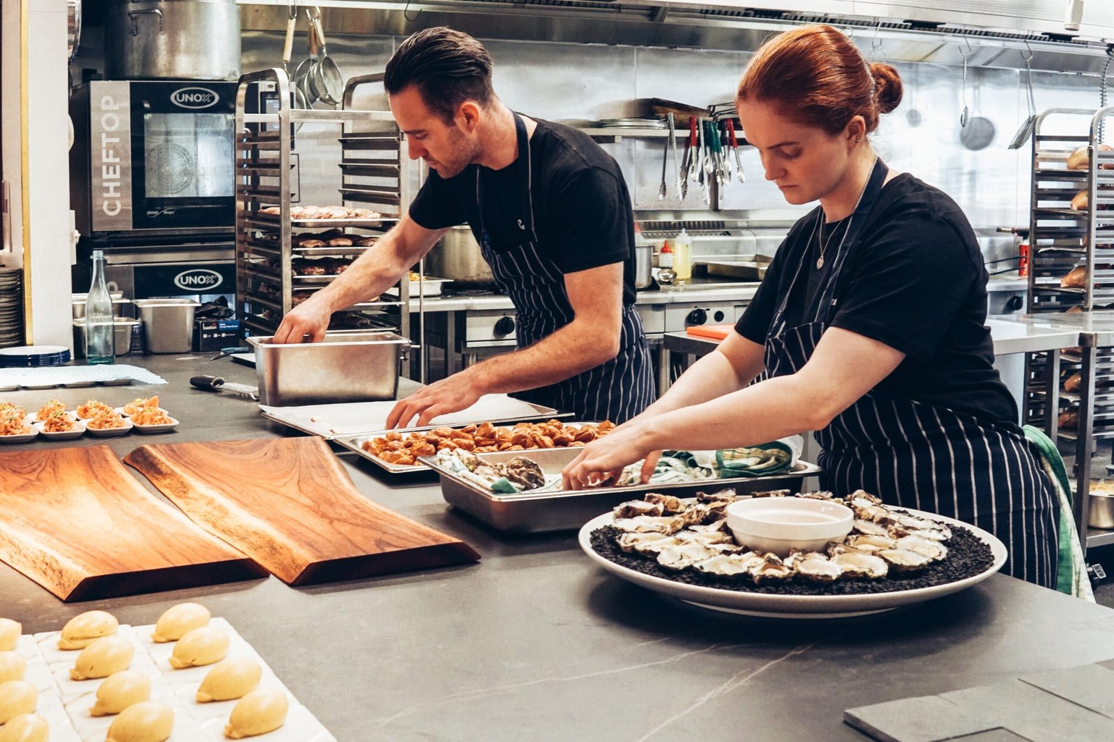 Two chefs, a man and a woman, both wearing black t-shirts and striped aprons, are actively preparing food on a stainless steel counter in a professional kitchen. The woman is arranging oysters on a large platter, while the man is working with other food items in metal trays.