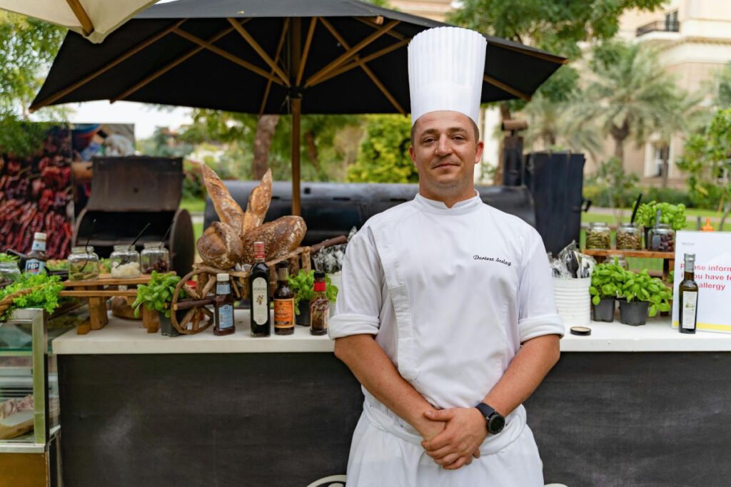 A male chef, wearing a white uniform and tall toque, stands smiling in front of an outdoor culinary setup featuring various ingredients, breads, and bottles under an umbrella at a resort.