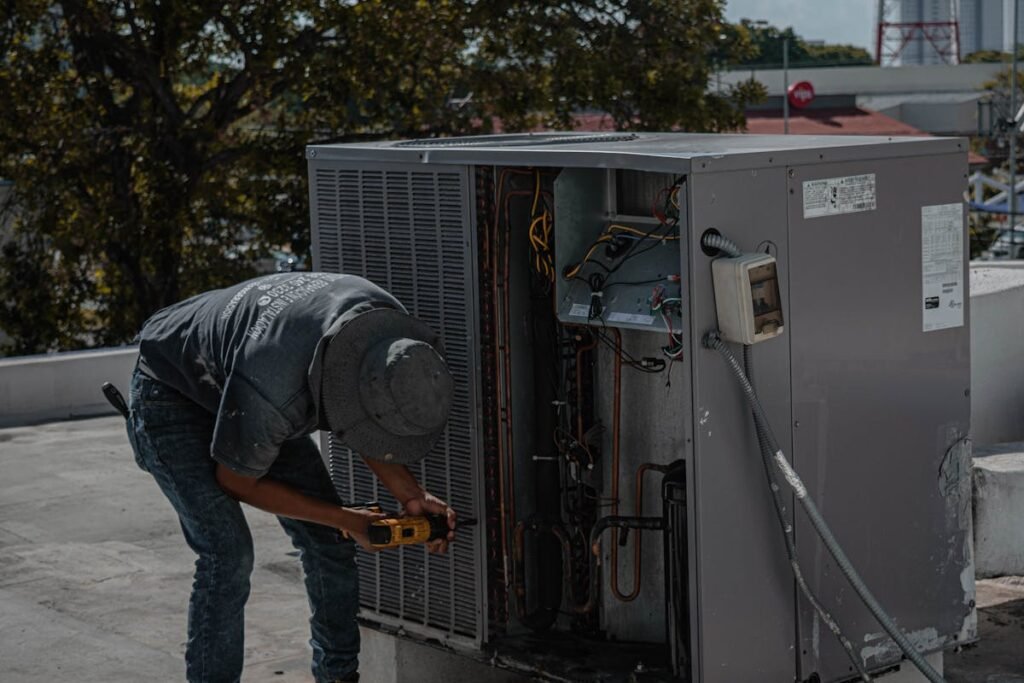 An HVAC technician wearing a hat and work clothes is bending over, using a power drill to work on the side panel of a large outdoor air conditioning unit on a rooftop.