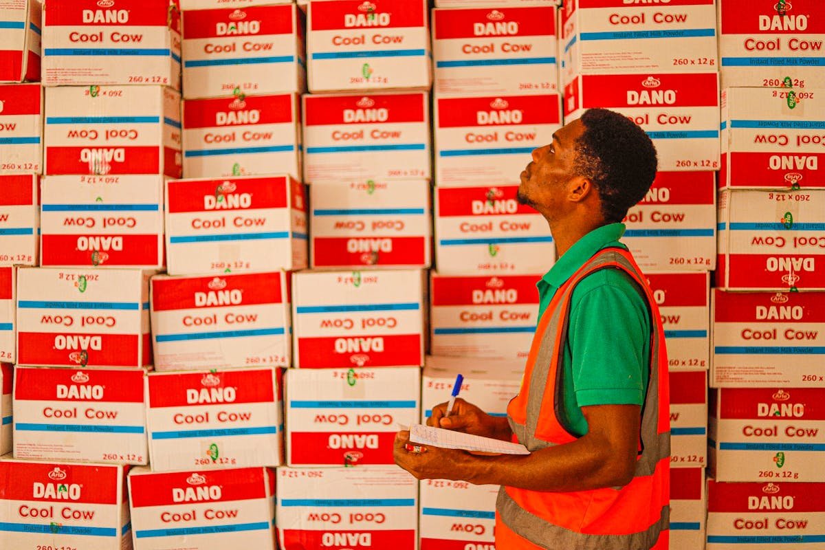 A male warehouse worker in a green shirt and orange safety vest looks up while writing on a clipboard, standing in front of tall stacks of red and white boxes labeled "DANO Cool Cow."