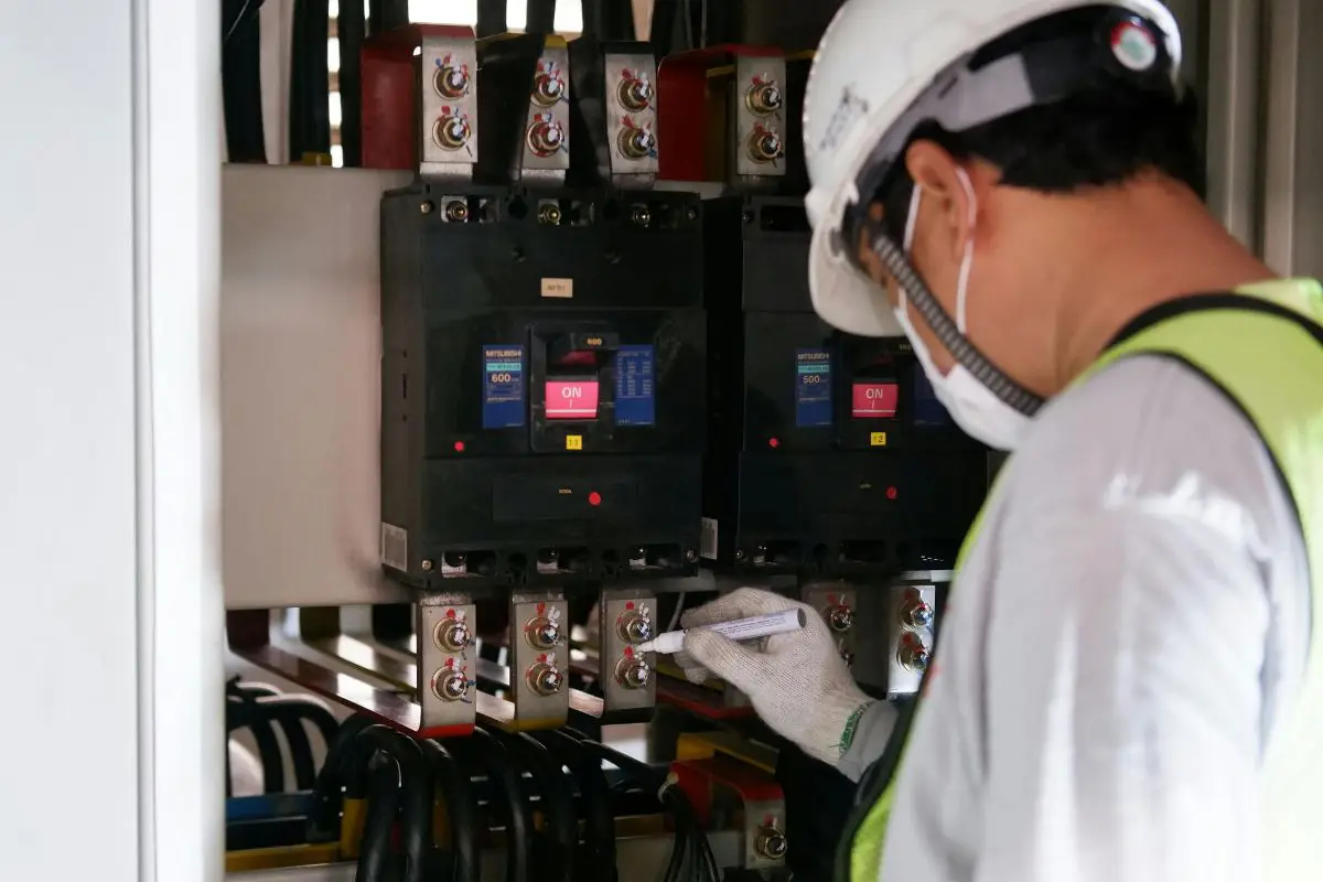 A technician in a hard hat and mask inspecting an open electrical panel with circuit breakers.