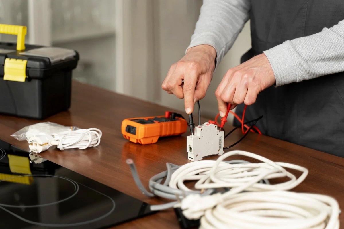 Close-up of an electrician's hands connecting wires to an electrical component with a multimeter, toolbox, and coils of wire on a wooden surface.