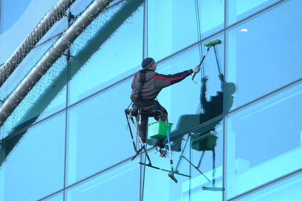 A lone window cleaner suspended by ropes on the side of a modern glass skyscraper.