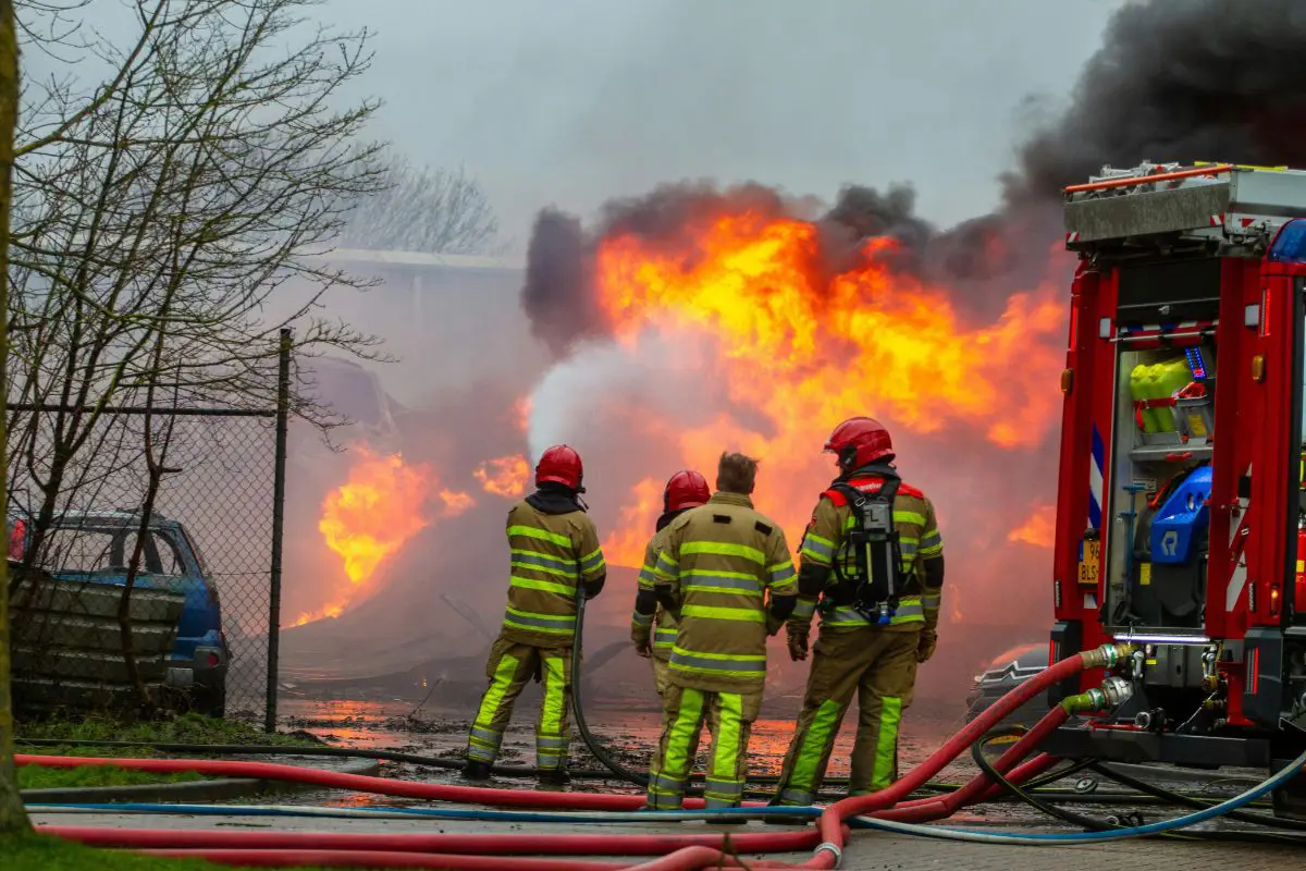 Firefighters in protective gear are spraying water at a large, uncontrolled fire with thick smoke.