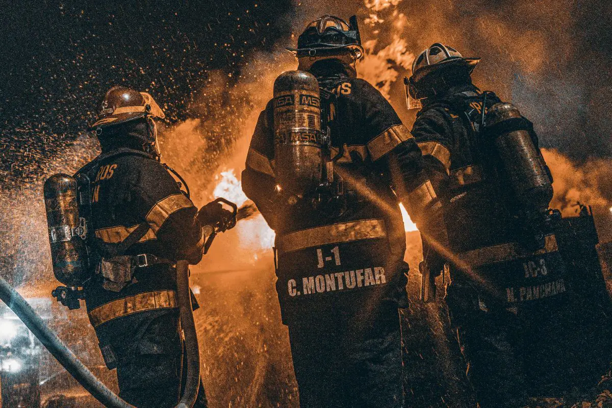 Three firefighters in full gear with air tanks on their backs, facing a large blaze in the dark.