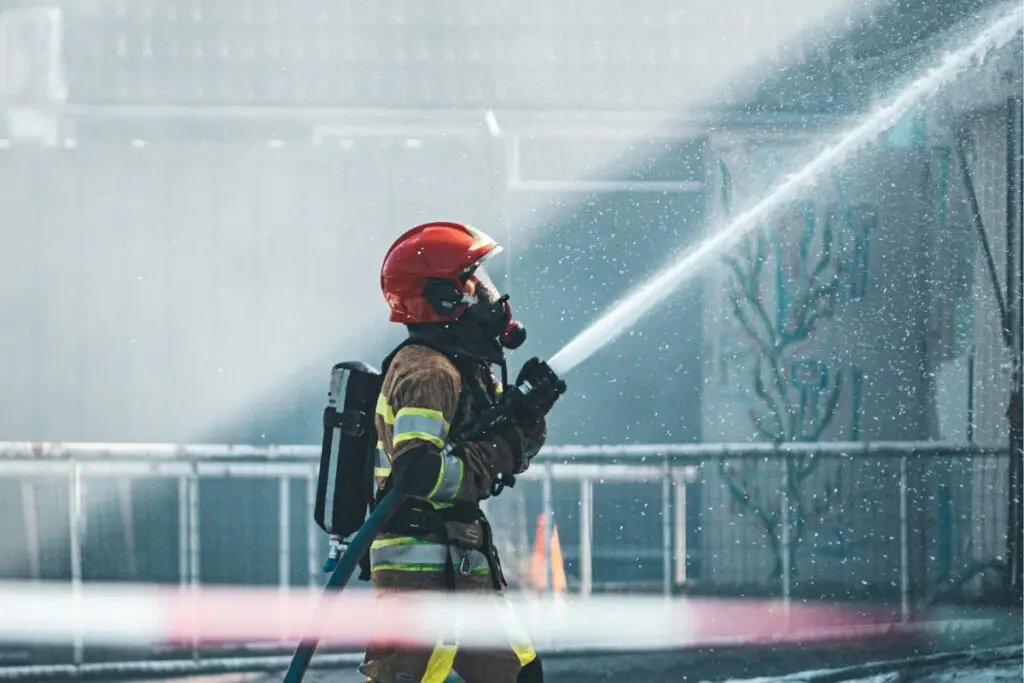 A firefighter in a red helmet and breathing apparatus directs a powerful stream of water.
