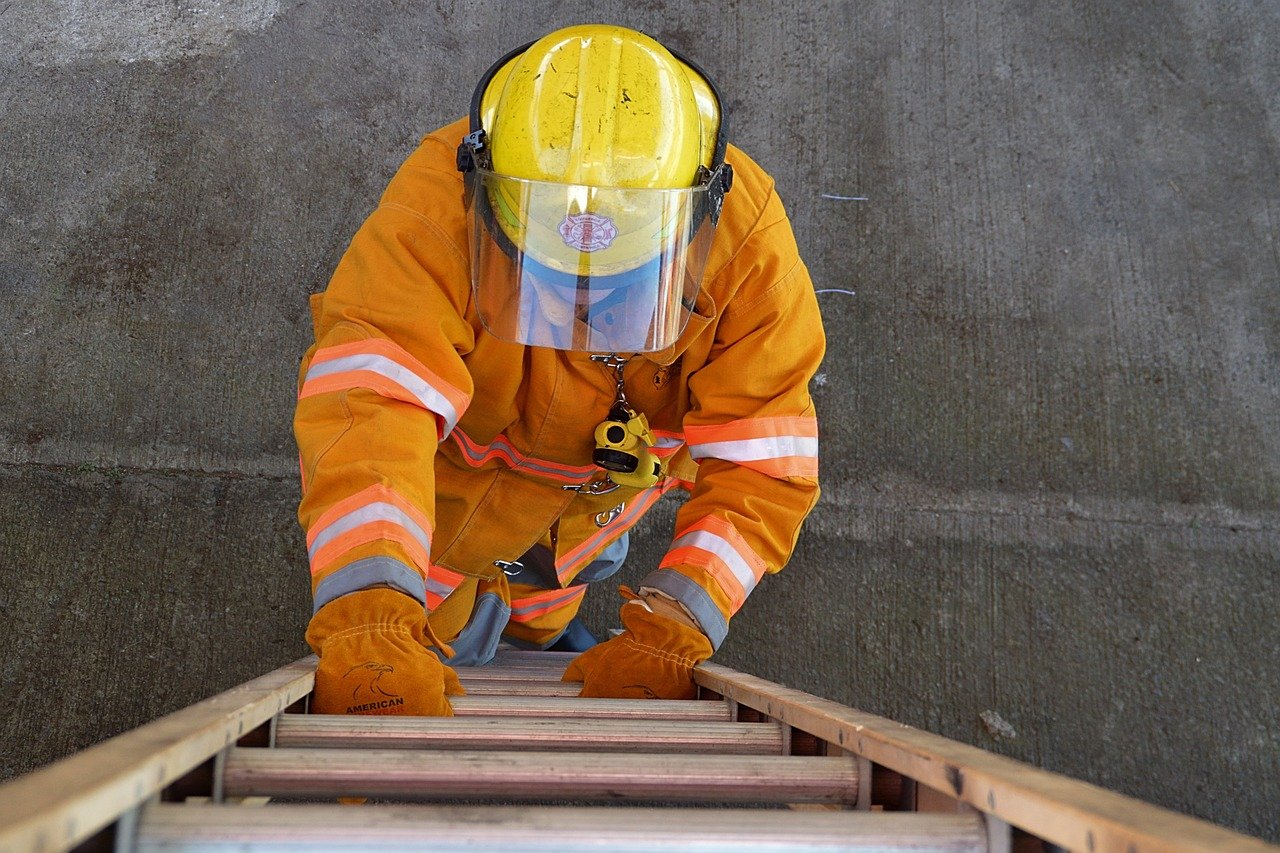 A firefighter in full orange protective gear, including a yellow helmet with a clear visor, is seen from above climbing a wooden ladder.