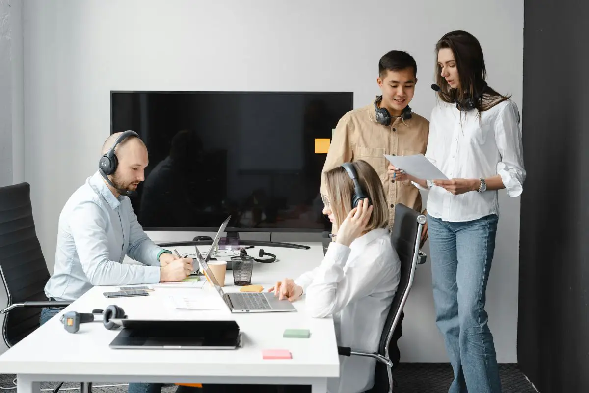Four professionals with headphones working at a modern office desk with laptops.