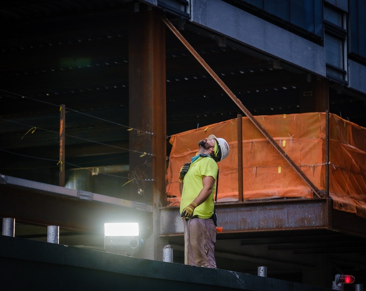 A construction worker in a hard hat, yellow shirt, and work gloves stands on a building structure, looking up intently with scaffolding and steel beams in the background.