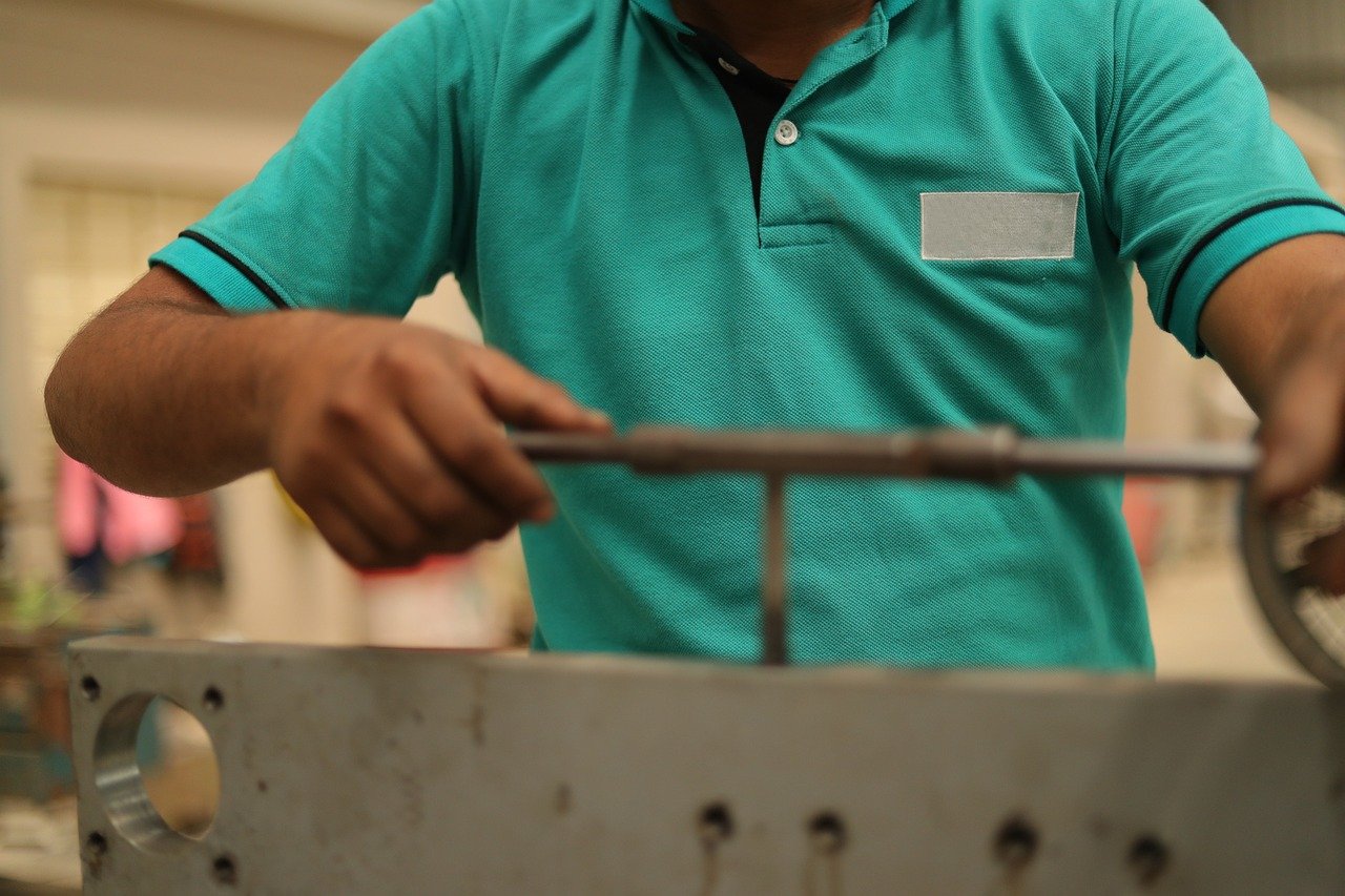A construction worker wearing a hard hat, safety glasses, and a high-visibility vest is measuring a metal frame for a ceiling installation with a tape measure, arms raised.