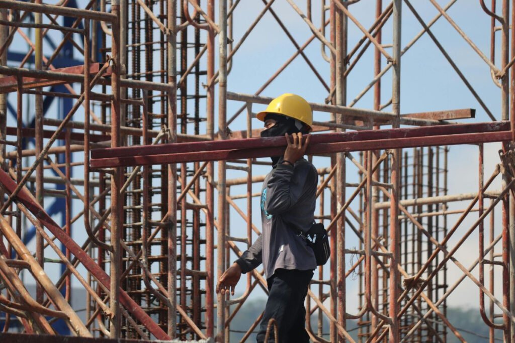 A construction worker in a yellow hard hat carries metal beams on their shoulder amidst scaffolding.