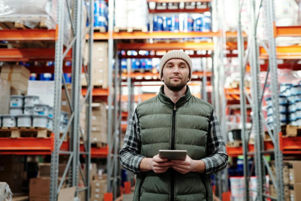 man in a beanie and vest holds a tablet in a warehouse aisle.