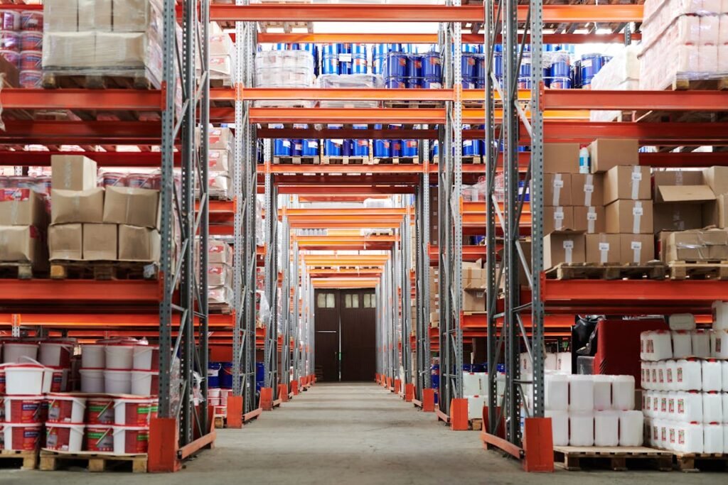 A long, narrow aisle in a warehouse lined with tall, orange metal shelving units packed tightly with boxes, barrels, and various packaged goods.
