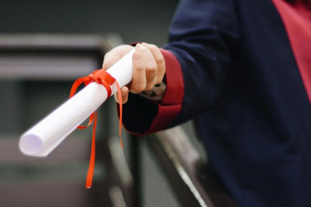 A hand holds a white rolled diploma tied with a red ribbon, wearing a graduation gown.