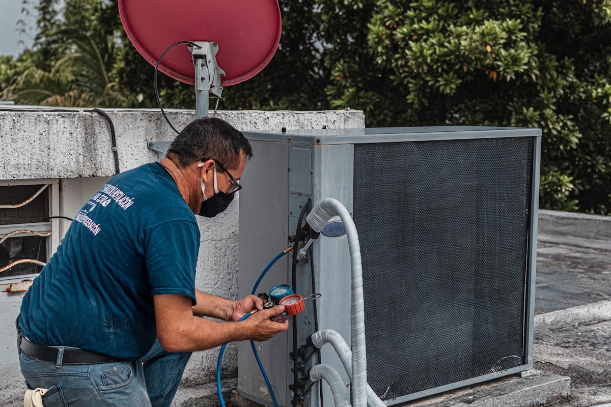 A technician wearing a blue shirt, face mask, and glasses is connecting gauges to an outdoor air conditioning unit on a rooftop, with a red satellite dish and green trees in the background.